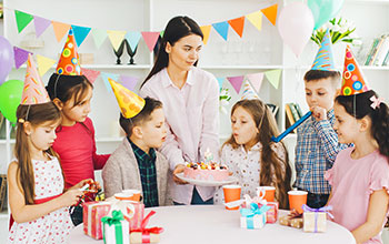 Mother holding a cake and kid blowing out candles.  5 other kids wearing party hats around the cake watching.
