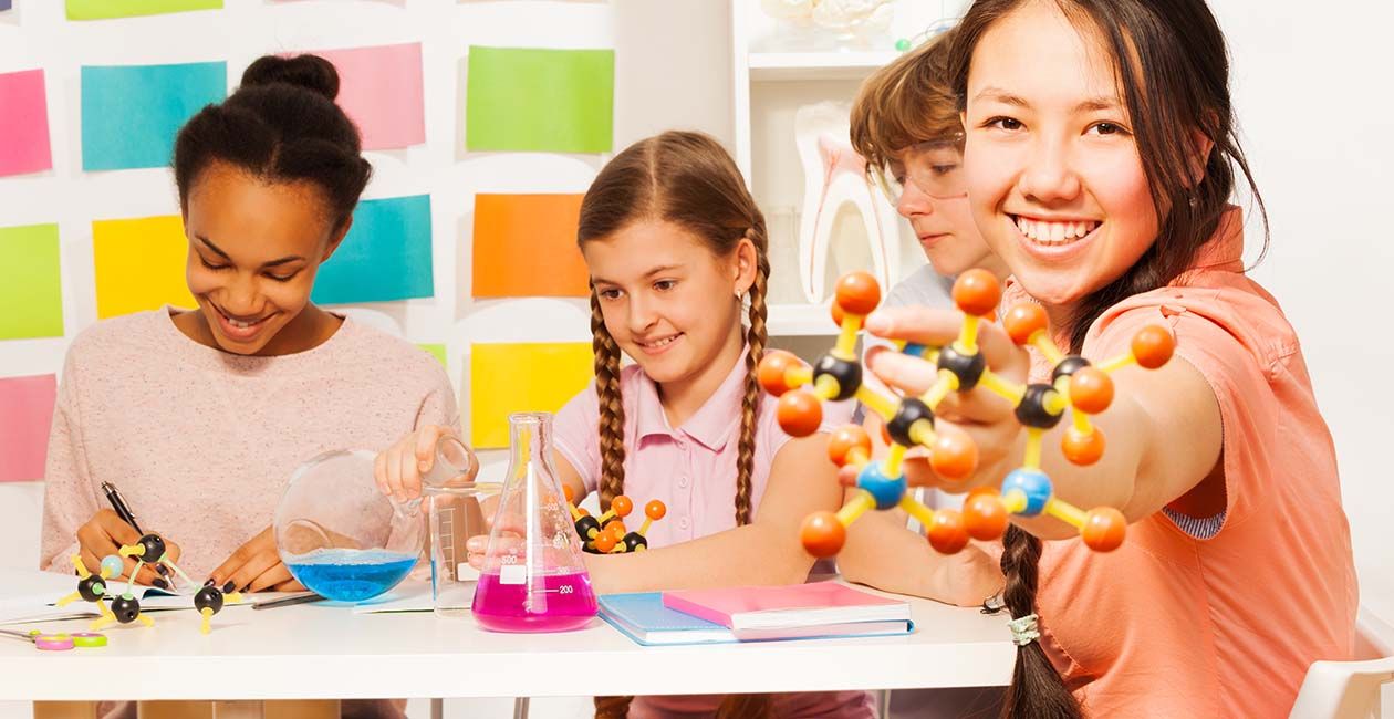 4 girls designing 3D atoms at desk with 2 beakers one blue one pink on the table